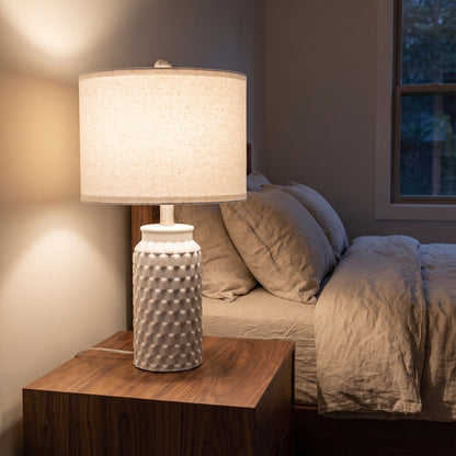 Ceramic table lamp glowing on a walnut nightstand beside a bed in the evening