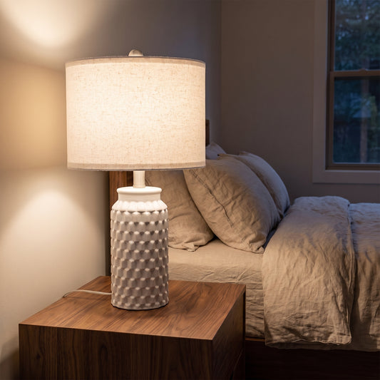 Ceramic table lamp glowing on a walnut nightstand beside a bed in the evening