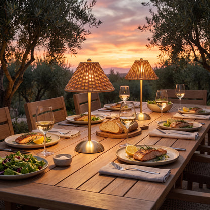 Two cordless rattan-look lamps lighting an outdoor dinner table at sunset